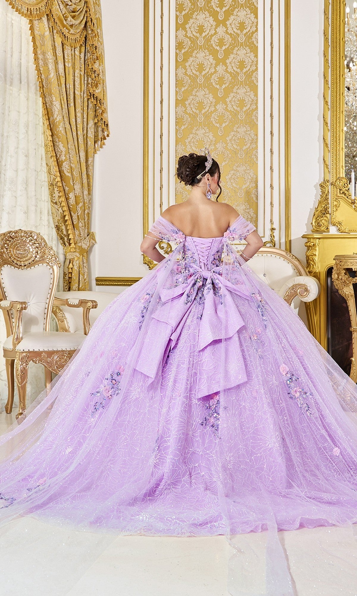 A woman stands with her back to the camera in an ornate, gold-accented room, wearing the Quinceanera Dress 5127 By Dancing Queen—a voluminous lavender ball gown with large bows and beaded floral details.